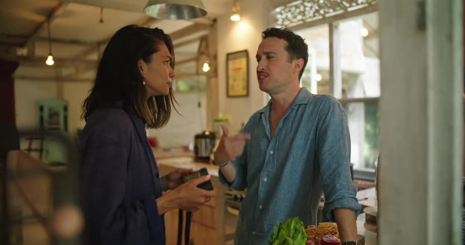 A man feeds a woman a tomato in their kitchen and the man makes an excited gesture like he just got something he wanted