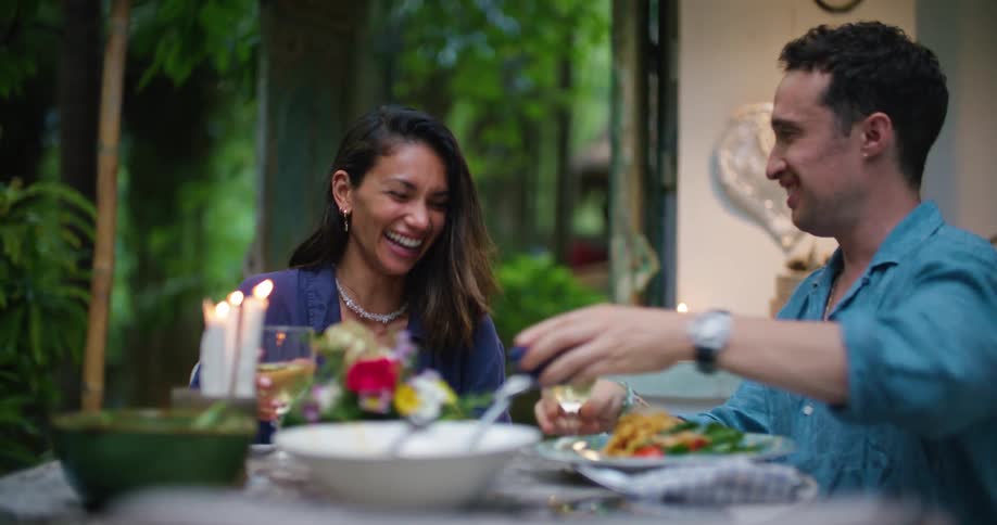 A man and a woman are sitting down at a table eating and talking and they raise their wine glasses and toast a drink