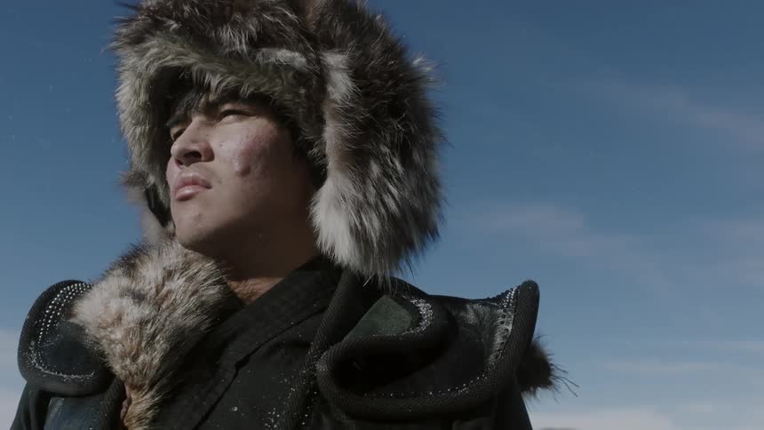 A man wearing a fur hat stands outside on a sunny day
