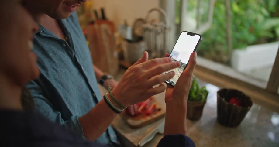 Man and woman standing in front of kitchen counter looking through pictures on the smartphone while talking and laughing