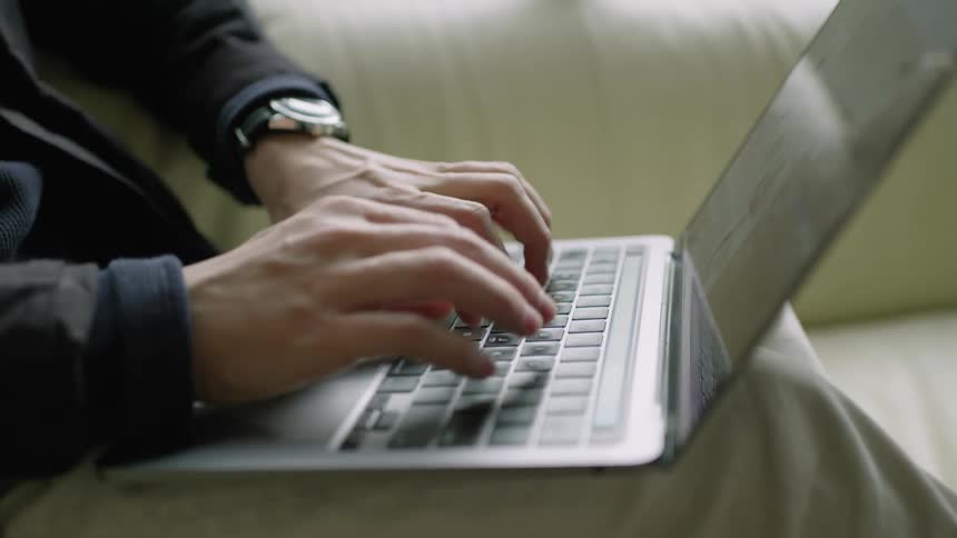 Close up of the hands of a man typing on a laptop sitting on a couch