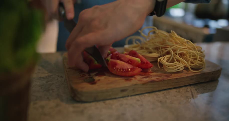 A man is slicing a tomato on a cutting board with spaghetti noodles on the side