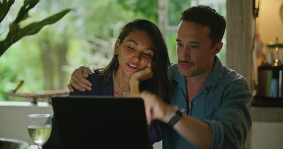 A man and a woman are sitting together at a table with a laptop computer talking and he has his arm around her