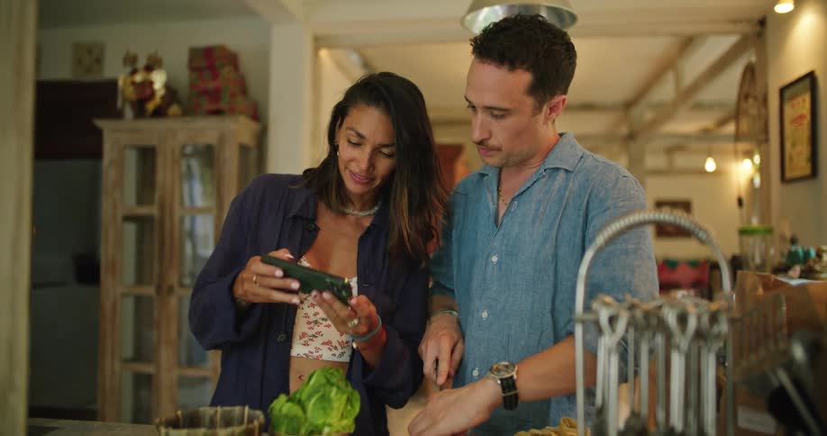 Woman shows pictures to his husband on her phone while he is cooking at home