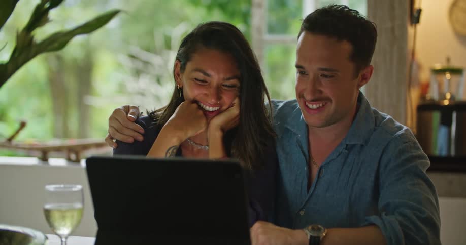Young happy couple hug while sitting at the dinner table looking at a laptop