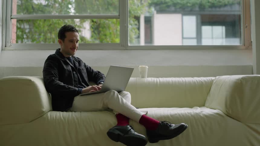 Young man sitting on a couch at home near a window typing on a laptop at daytime