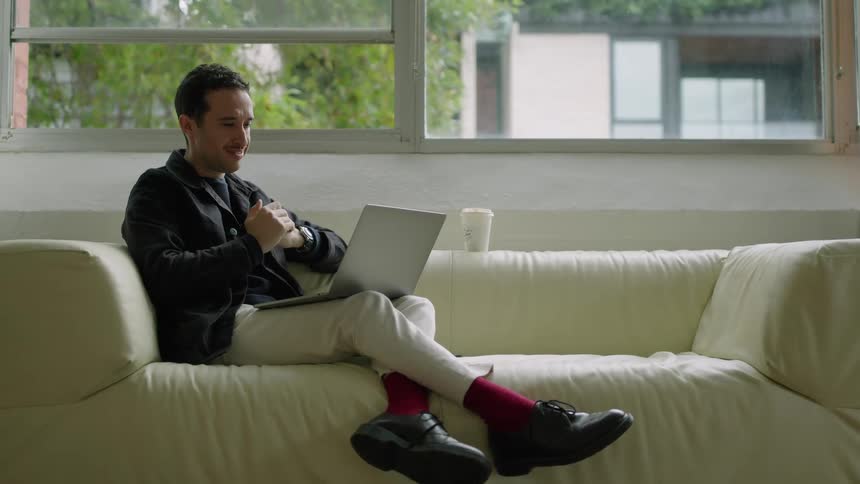 Man sitting on a couch at home talks to himself while working on a laptop