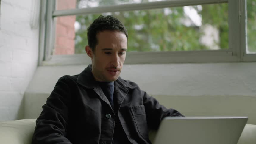 A man sits on a couch talking to someone on his laptop while sipping out of a mug