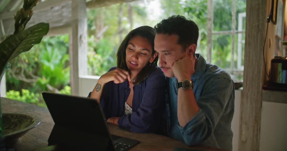 A man and woman sit at a kitchen table working diligently using a touchscreen laptop and a smartphone