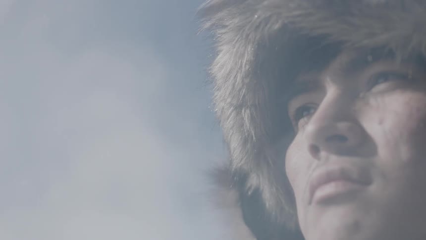 A man wearing a fur hat stares ahead while mists sweep across his face