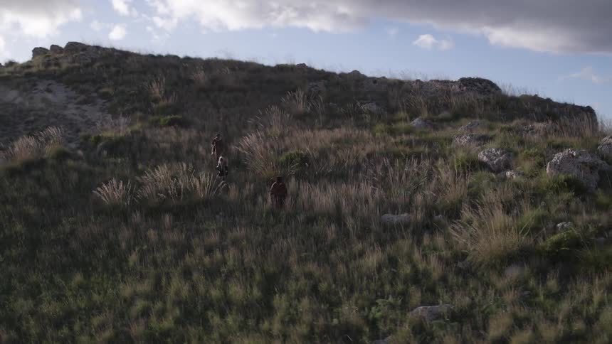 Three people in long robes hike down a hillside during the day