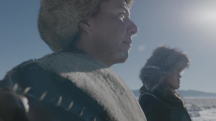 Two men in heavy clothing and fur hats stand on a snow covered field on a cold day