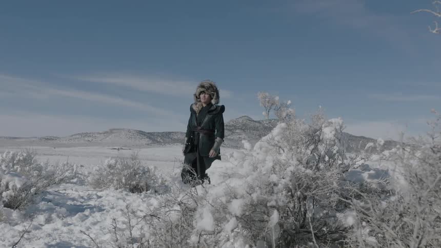 A man in heavy clothing and a fur hat walks through the snow on a cold sunny day