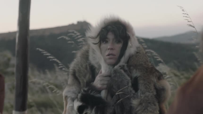 A woman wrapped in fur pelts looks at a person standing behind her in a stunned manner
