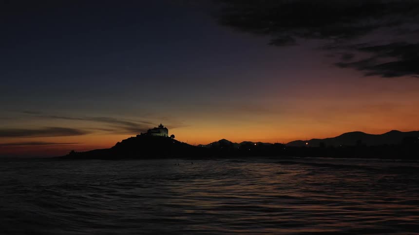 The silhouette of hills and church along a coastline are illuminated by the sunset behind them