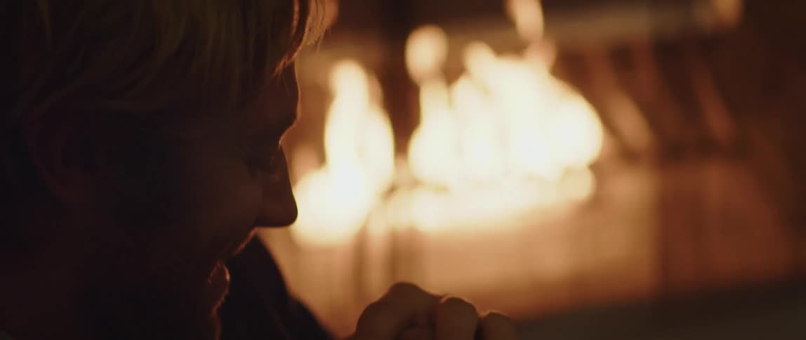 A man smiles and bows his head while sitting outside near a flaming fire place