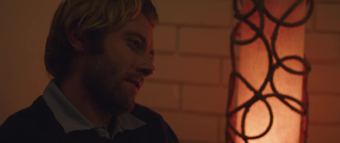 A man smiles and smirks while sitting in a dim room