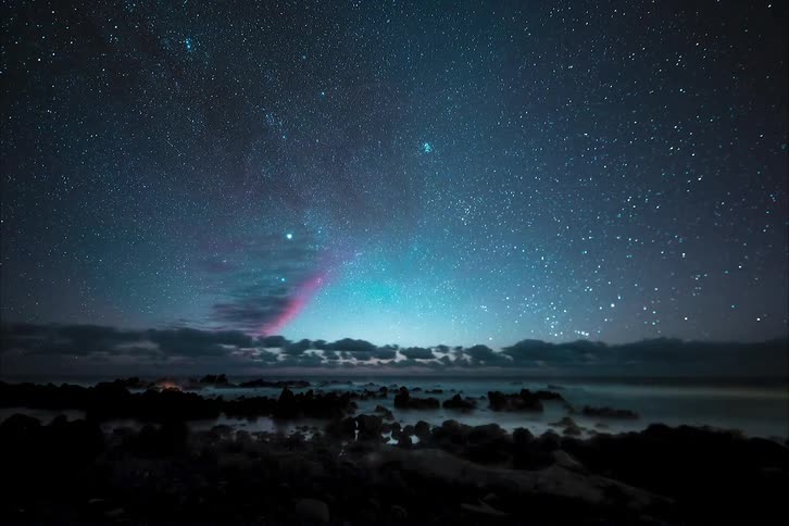 Stars of the milky way move across the sky with shooting stars over the water along the shore and then clouds are visible as the sun rises