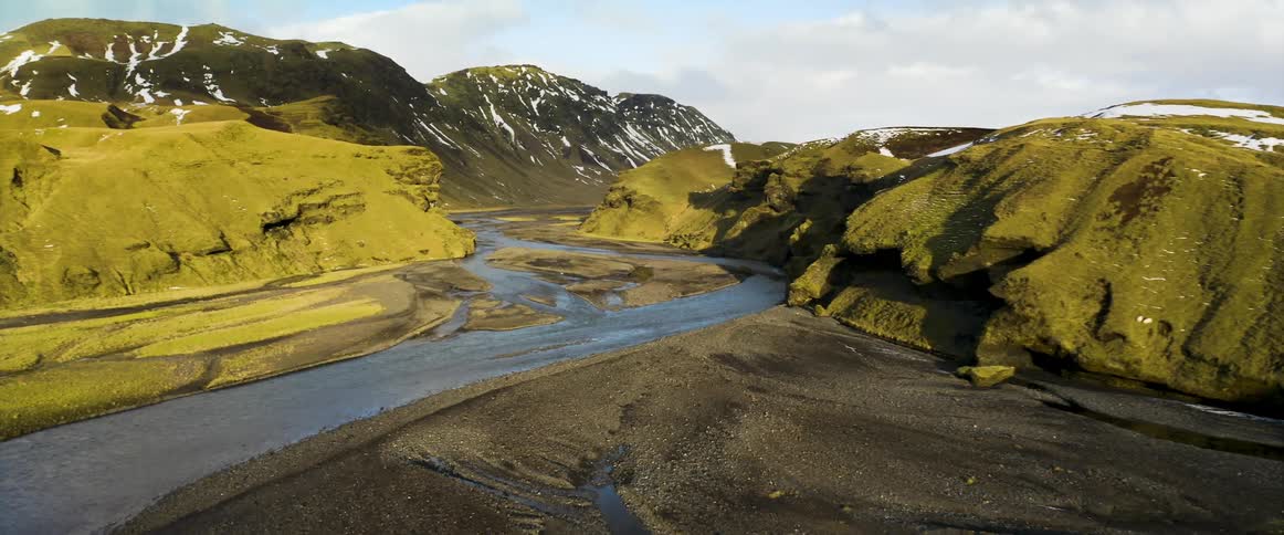 A river runs through a canyon in a mountain range that still has snow on the peaks