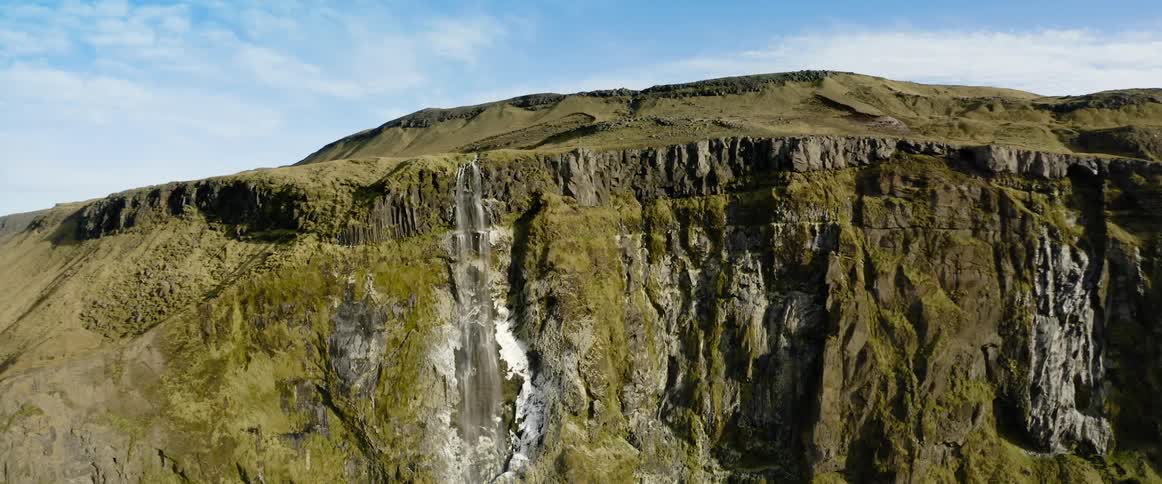 A waterfall down the cliff of a mountain covered in short vegetation during the day
