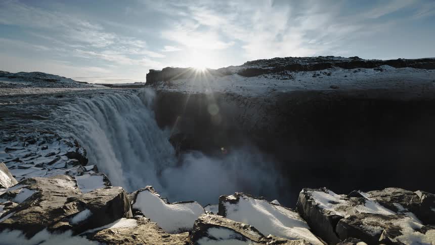 Sun shines over the jagged edge of a mountain next to a waterfall on a snowy landscape