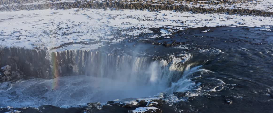 A riverbank dusted with snow as water rushes over in a waterfall