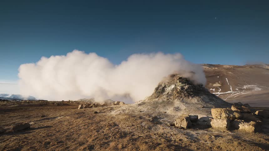 Thick clouds of steam coming out of a vent in the ground next to the mountains under a clear blue sky