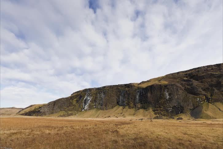 Thin waterfall falls through crevice cut into face of cliff bordering field on cloudy day