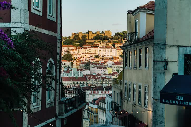 Pedestrians move along street at top of slope with fortification on top of hill across town