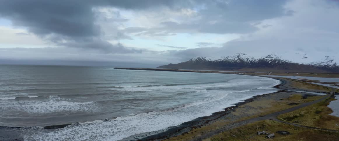Aerial view of waves gently crashing on the coast of a beach close to a snow covered mountain range at evening