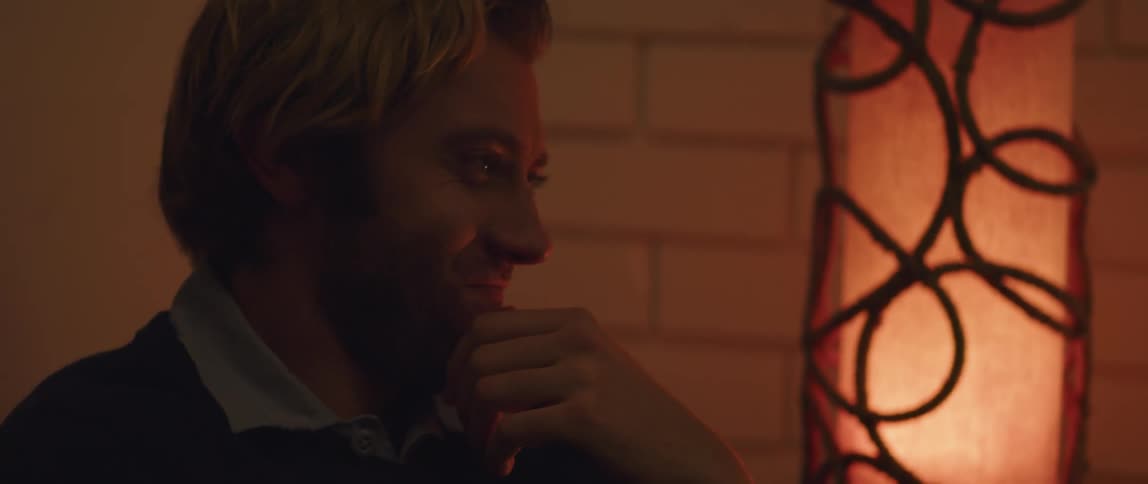 Close up of the face of a man sitting on a dimly lit room covering a smile with his hand