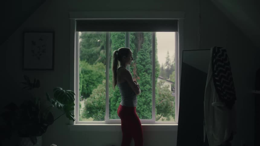 A lonely woman standing beside a window in front of a mirror with her arms crossed in a dark room
