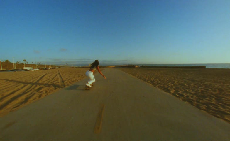 Woman with cap yellow top and white pants skateboards on road on the beach