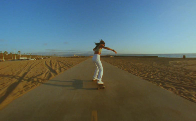 Woman with cap yellow top and white pants rides skateboard on road on the beach doing tricks