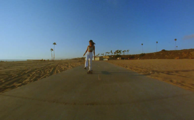 Woman with cap yellow top and white pants skateboards on road on the beach