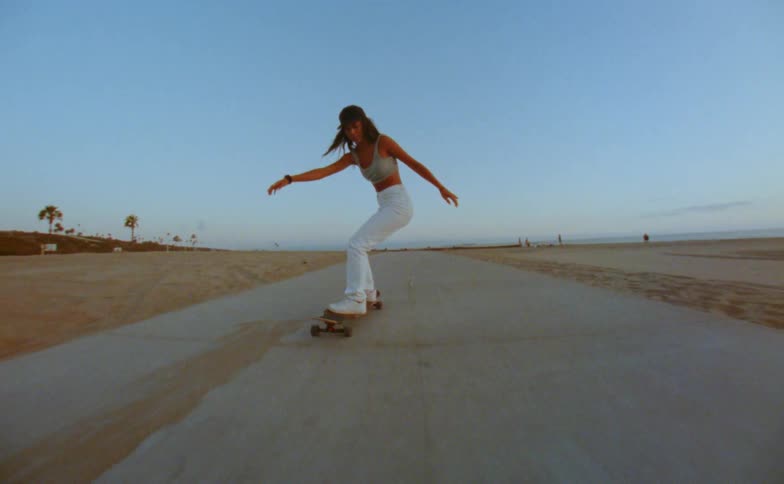 A teenage girl raises her arms in the air as she rides her skateboard on a road near the beach