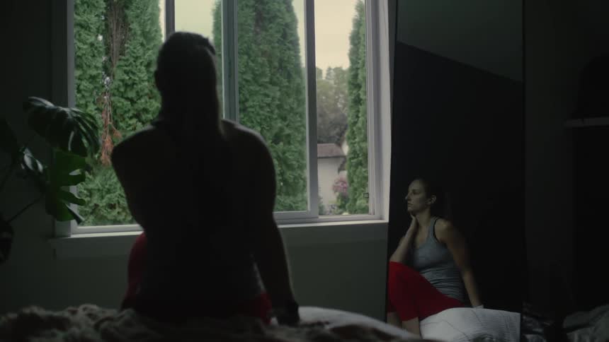 Two women sitting on a dimly lit room over pillows look out the window of a bedroom