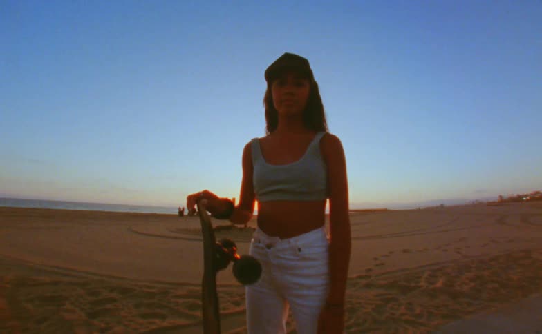 Young woman stands with a long skateboard on her hand near the beach at sunset