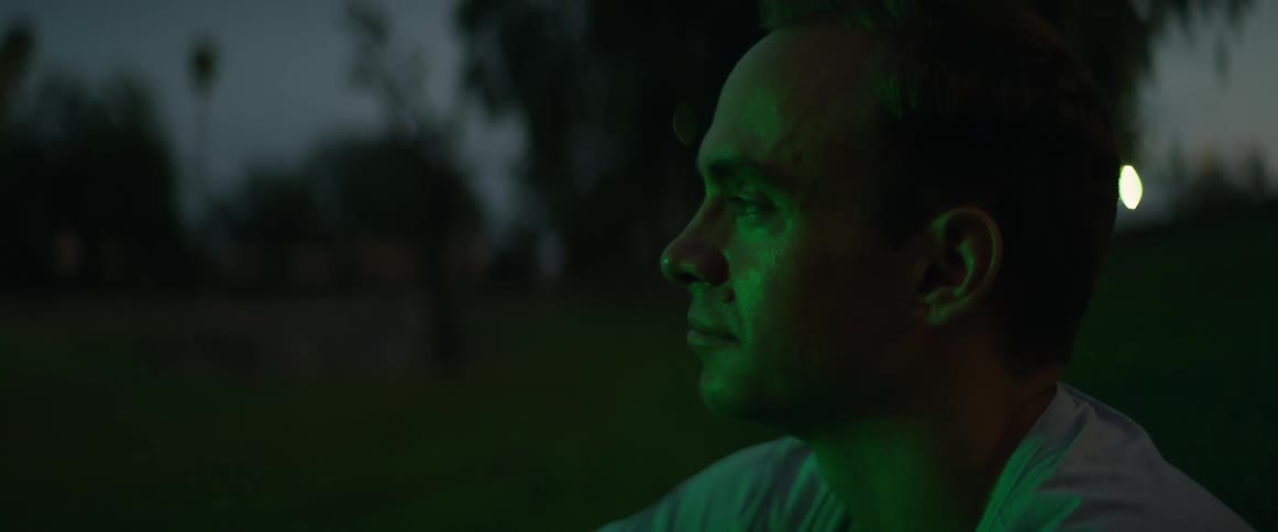 A man sits outside in the grass watching something with flashing lights and seems to be deep in thought