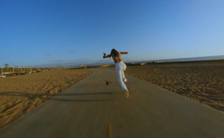 Woman with cap yellow top and white pants skateboards on road on the beach doing tricks