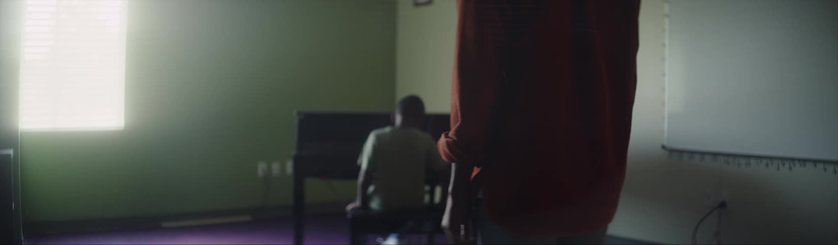 Father standing in the middle of a room while his son walks to a piano sits down and starts playing