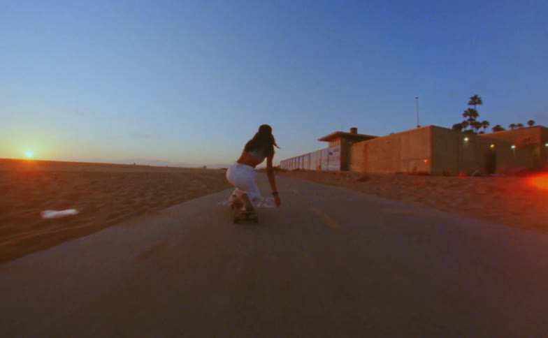 Woman crouches on a skateboard and then stands up while going down a road near a beach