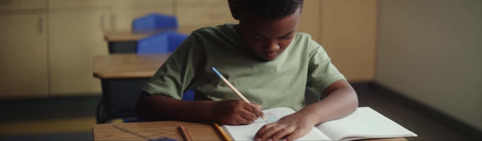 Child sitting on a desk in the classroom drawing with colored pencils on his notebook