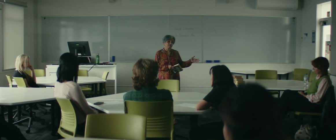 Woman standing in the middle of a classroom holding a book and talking to women sitting on the tables listening to her