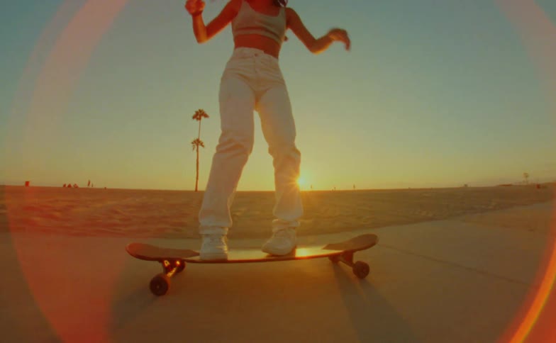 Sun sitting near the horizon causes camera glare as female skateboarder rides down sidewalk