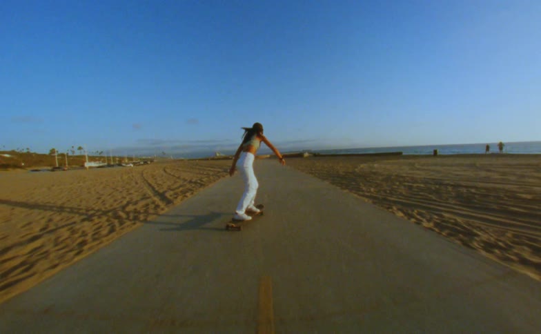A teenage girl raises her arms in the air as she rides her skateboard on a road near the beach