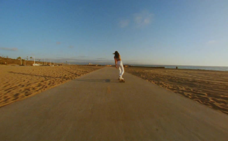 Woman with cap yellow top and white pants skateboards on road on the beach