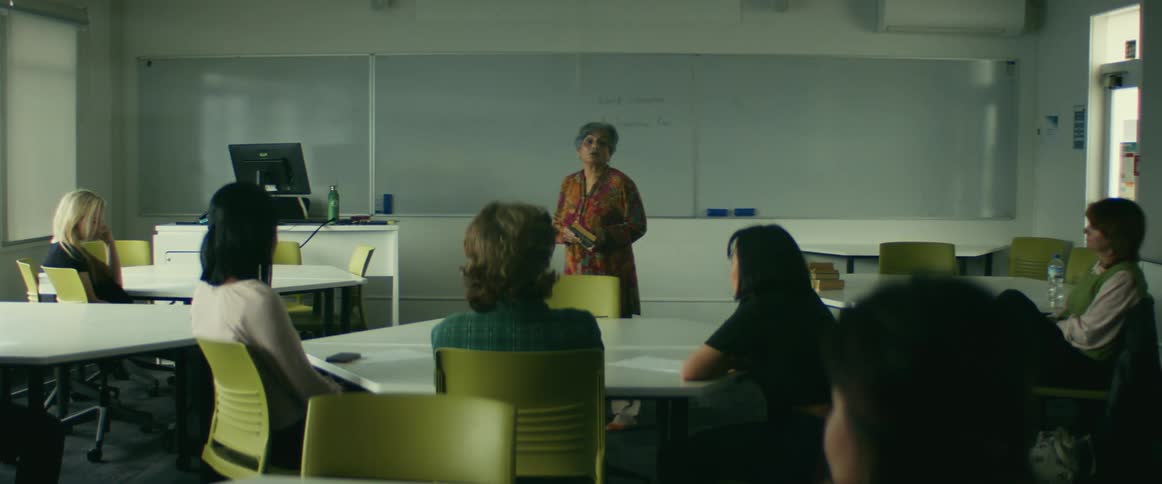 Woman holding a book in the middle of a classroom in front of the whiteboard giving class to some adults