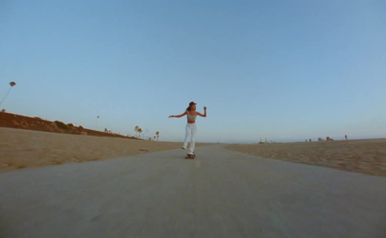 A teenage girl raises her arms in the air as she rides her skateboard on a road near the beach