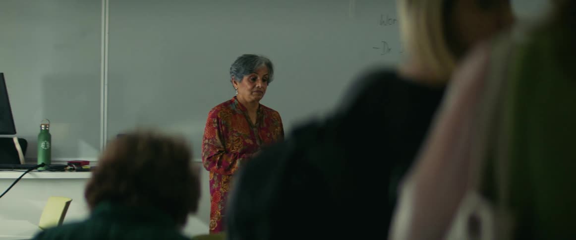 Instructor walks away from desk in classroom as students take their seats